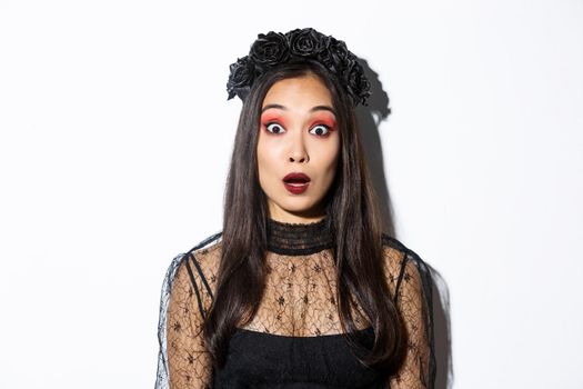 Close-up of impressed and shocked asian woman in halloween costume, drop jaw and gasping wondered, standing over white background.