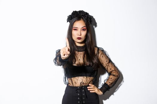 Serious-looking sexy asian woman in black gothic dress celebrating halloween, showing stop gesture, shaking finger to prohibit or refuse something, standing over white background.