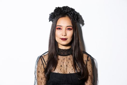 Close-up of beautiful gothic girl with black wreath, getting dressed for halloween party, standing over white background in wicked witch costume.