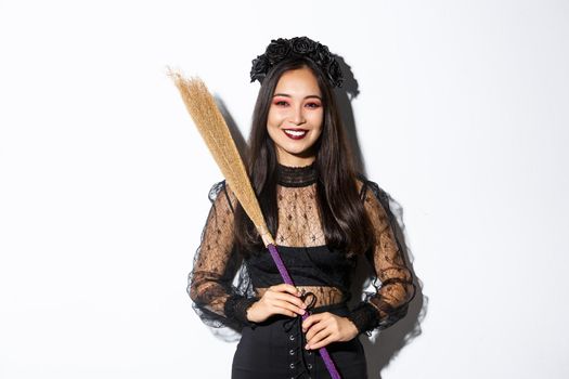 Portrait of smiling beautiful asian woman in witch costume holding broom and looking happy at camera, celebrating halloween, enjoying trick or treating, white background.
