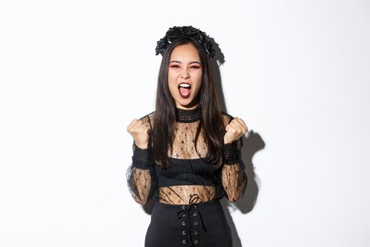 Image of joyful attractive asian witch, female in halloween costume yelling in rejoice and fist pump, celebrating victory, triumphing over white background.