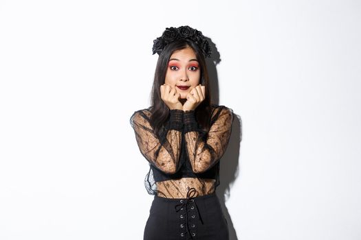 Excited cute asian girl in witch costume looking hopeful at camera, daydreaming while standing over white background, wearing gothic lace dress with wreath.