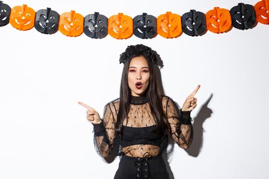 Joyful asian woman in gothic lace dress celebrating halloween, pointing fingers sideways, standing against white background with pumpkin party streamers.