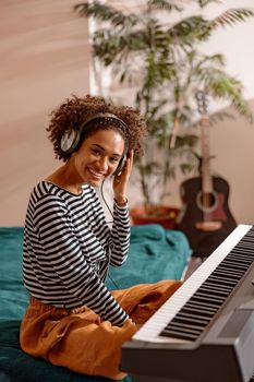 Joyful multiracial female musician looking at camera and smiling while sitting on bed in front of electronic musical instrument