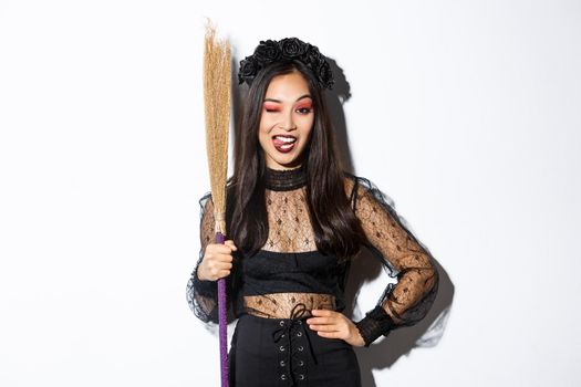 Sassy good-looking asian woman in witch costume showing tongue, holding broom and posing over white background.