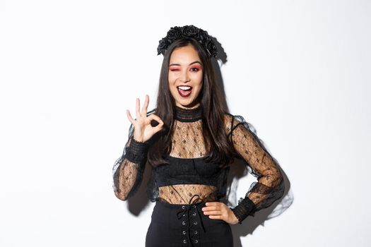 Attractive young asian woman in halloween gothic dress, showing okay gesture, give permission, approve or like your party costume, standing pleased over white background.