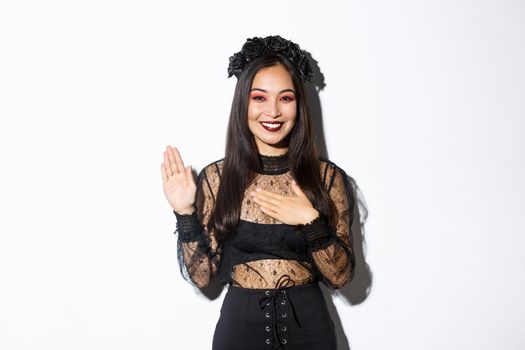 Image of cute sincere asian girl in halloween costume making promise, holding one hand on heart while swearing, being honest, standing over white background.