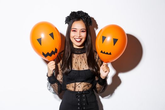 Image of cheerful asian woman in witch costume celebrating halloween, holding balloons with scary faces, standing over white background.