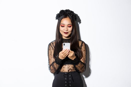 Image of stylish asian woman in halloween costume checking messages on mobile phone. Girl in gothic lace dress looking at smartphone, standing over white background.