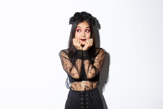 Silly beautiful asian girl in gothic lace dress with wreath looking dreamy left, have nostalgic thoughts, standing in halloween costume over white background.