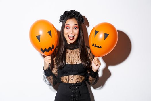 Image of happy asian woman in witch costume celebrating halloween, holding balloons with scary faces, standing over white background.