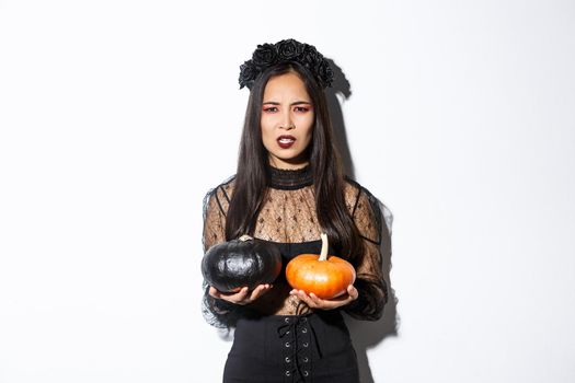 Image of grimacing angry witch throwing pumpkins, girl celebrating halloween, standing over white background.