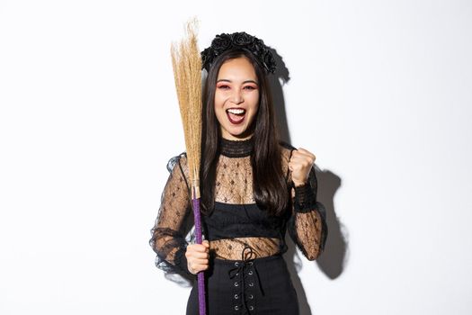 Image of cheerful asian girl in witch costume celebrating victory, holding broom, saying yes and raising fist in triumph, white background.