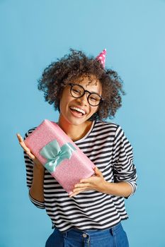 Happy joyful female person in birthday cone hat looking at camera and laughing while holding gift box. Isolated on blue background