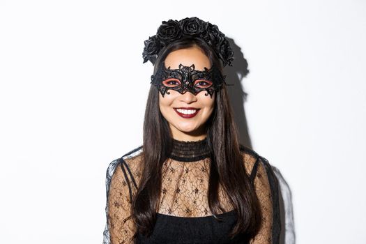 Close-up of mysterious woman in gothic wreath and black mask smiling at camera, celebrating halloween, standing over white background.