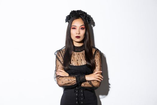 Skeptical young asian woman in witch or widow costume looking doubtful. Female dressed-up for halloween party, wearing black lace dress and wreath.