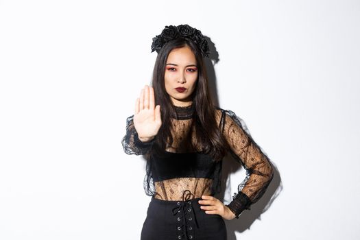 Image of serious asian woman in halloween costume of witch, showing stop gesture, forbid or prohibit something with displeased confident face, standing white background.