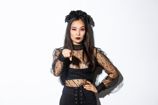 Arrogant and sassy asian evil witch, woman in halloween costume, pointing finger at you and looking mysterious, standing over white background.