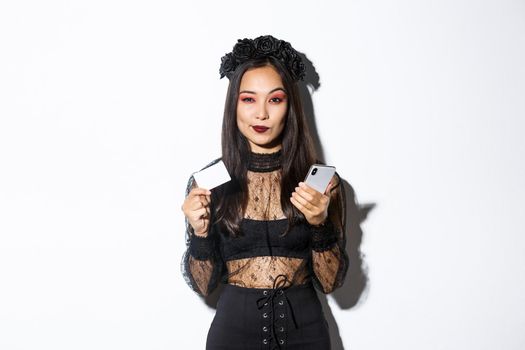 Sassy young woman looking thoughtful, holding credit card and mobile phone, shopping in internet, standing over white background.