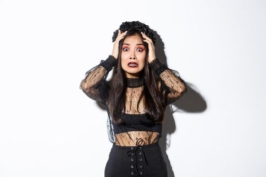 Image of scared and worried asian woman in halloween costume looking horrified or anxious, holding hands on head and stare at camera, panicking over white background.
