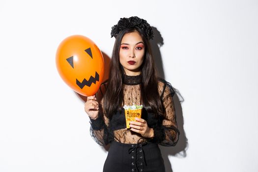 Serious-looking woman looking arrogant in witch costume, trick or treating during halloween night, holding balloon and sweets.