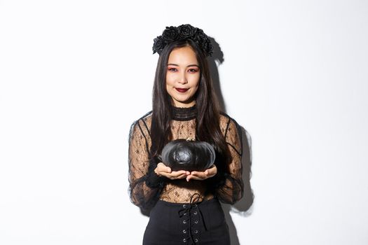 Smiling devious asian girl in witch costume, celebrating halloween, holding black pumpkin, standing over white background.