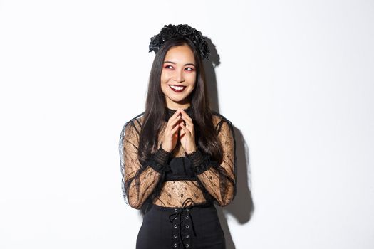 Image of beautiful asian woman dressed-up as a witch or widow in black lace dress, gothic wreath, smiling pleased upper left corner and steeple fingers while prepare plan.