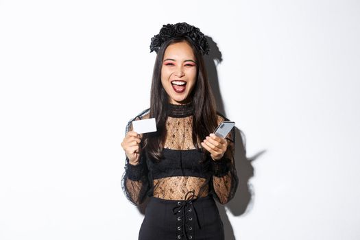 Happy laughing asian girl in halloween costume looking upbeat, holding smartphone and credit card, standing over white background.
