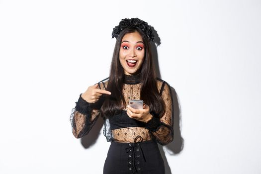 Portrait of excited asian girl pointing finger at smartphone, showing something about halloween in internet, standing over white background in gothic lace dress.