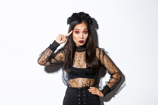 Image of annoyed young woman in halloween gothic dress scolding someone stupid, rolling finger on temple and looking at crazy person, standing over white background.