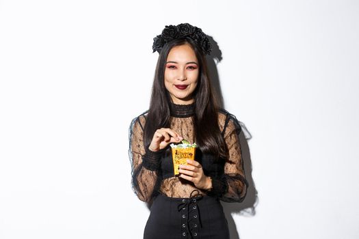 Smiling cute asian woman celebrating halloween, holding sweets and grinning happy, trick or treating in witch costume.