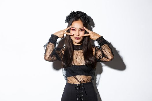 Beautiful asian woman with gothic makeup, wearing black widow dress and showing peace gesture, celebrating halloween, standing over white background.