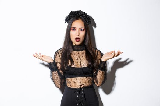 Confused asian woman in halloween costume looking clueless, raising hands sideways and stare puzzled at camera. Female wearing black lace dress, being a witch or magician on trick or treat party.