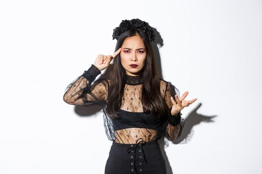 Frustrated asian woman in halloween costume arguing and complaining, scolding for something stupid, pointing finger at head and grimacing, standing over white background.