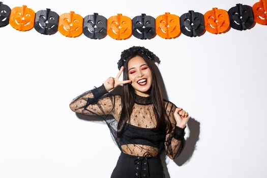 Carefree smiling asian woman in witch costume enjoying halloween party, dancing and showing peace gesture, standing over white background with pumpkin decoration.