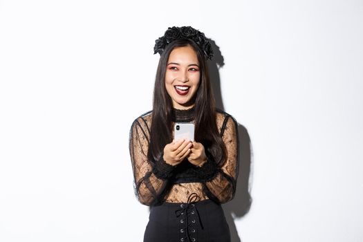 Happy attractive asian woman in halloween costume laughing carefree, holding smartphone and looking amused at upper left corner, standing over white background.