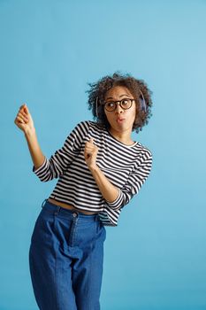 Lady in headphones enjoying favorite songs and making funny face while standing in studio. Isolated on blue background