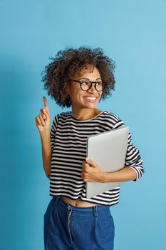 Cheerful multiracial lady looking away and smiling while holding notebook. Isolated on blue background