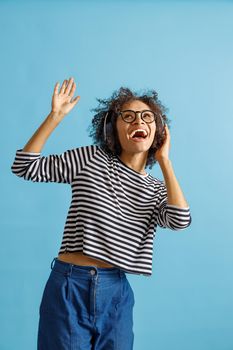 Joyful multiracial lady looking up and laughing while listening to favorite songs through wireless headphones. Isolated on blue background