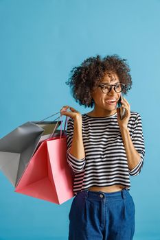 Joyful multiracial lady having phone conversation and smiling while holding purchases. Isolated on blue background