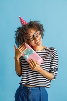 Cheerful multiethnic lady in party cone hat keeping eyes closed and smiling while holding gift box. Isolated on blue background