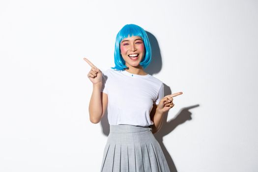 Joyful party girl in blue wig having fun, pointing fingers sideways, showing left and right promo, standing over white background.