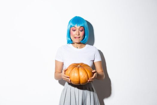 Image of cute girl picking pumpkin for halloween, wearing blue wig, standing over white background.