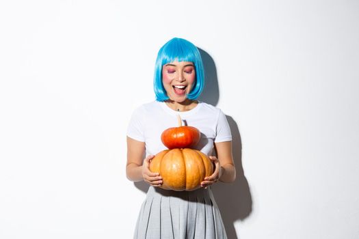 Happy asian girl in blue wig holding two cute pumpkins and smiling at camera, wearing schoolgirl outfit for halloween party.