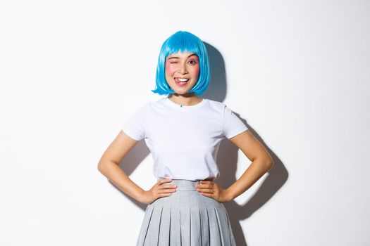 Portrait of beautiful asian girl in blue party wig showing tongue and winking silly, standing over white background in kawaii halloween costume.