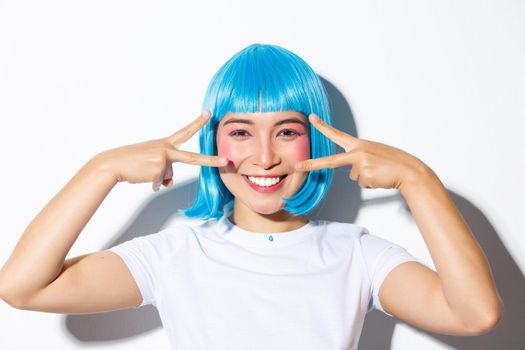 Image of cute asian woman in blue wig showing kawaii peace gesture and smiling, celebrating halloween, standing over white background.