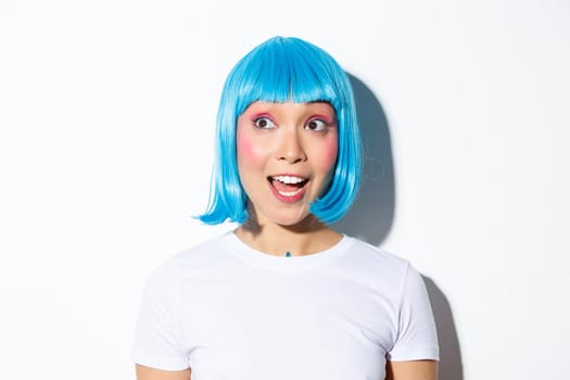 Close-up of surprised cute asian woman in blue wig, looking at halloween banner, standing over white background.