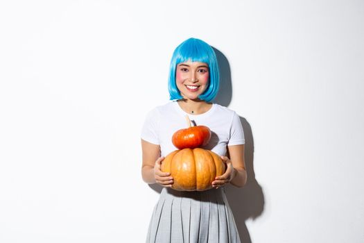 Adorable asian woman in blue wig holding two cute pumpkins and smiling at camera, wearing schoolgirl outfit for halloween party.
