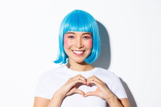 Close-up of beautiful smiling woman in blue wig, celebrating halloween, showing heart gesture, standing over white background.