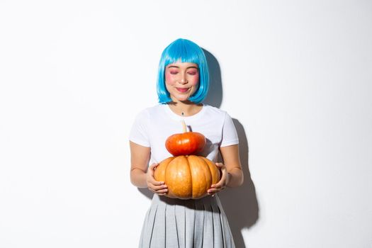 Dreamy cute asian girl standing with two pumpkins in blue wig and halloween costume, smiling with eyes closed.